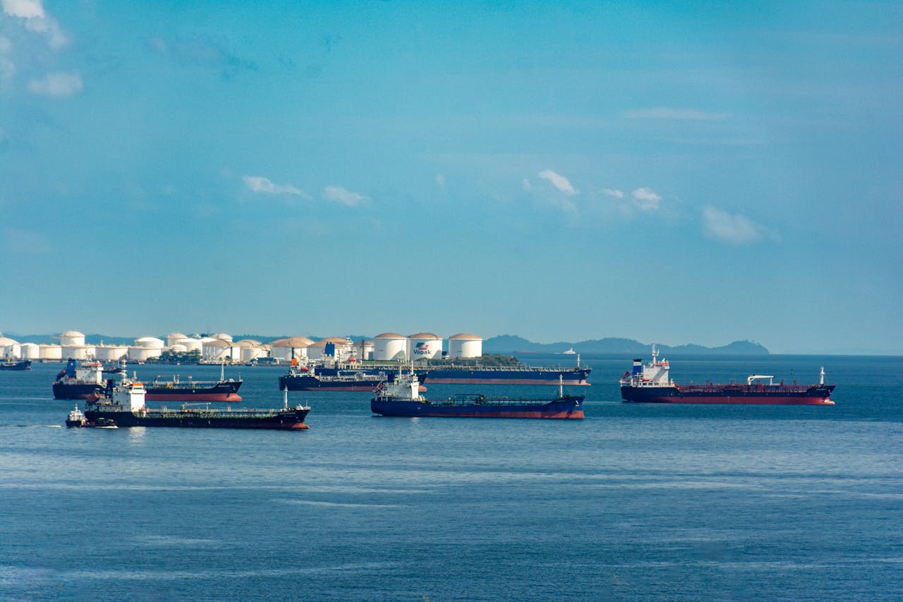 A fleet of cargo ships docked near oil storage tanks along a serene coastline with a clear blue sky above.