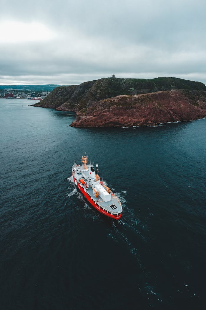 Services A dramatic aerial shot of a red cargo ship navigating near a rocky island under a cloudy sky.